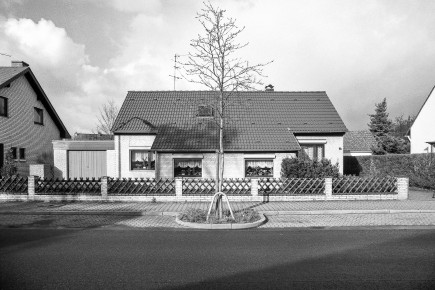 Im Vergleich zu den benachbarten Einfamilienhäusern neueren Datums werden die beschränkten Ausmaße der alten Siedlungshäuser besonders deutlich. Abgebildet ein Haus Typ Spiegel (Name des Architekten) in einer gespiegelten Version. Die Spiegelebene läuft senkrecht durch das Bild, der ursprüngliche Entwurf ist nur halb so groß. Kurz vor den Aufnahmen wurden die Straßen der Siedlung neu geteert. Die vormals unbefestigten Bürgersteige wurden auf dem Sandweg gepflastert und Parkbuchten eingerichtet.